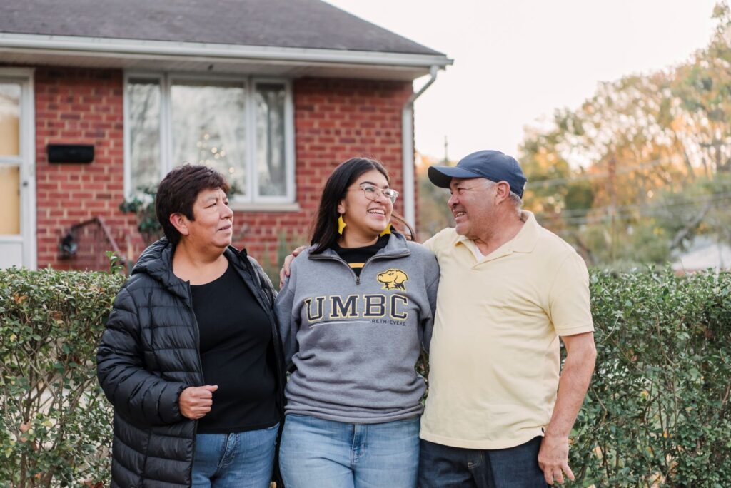 Young woman in UMBC sweatshirt smiles while embracing two older adults, standing in front of a home.